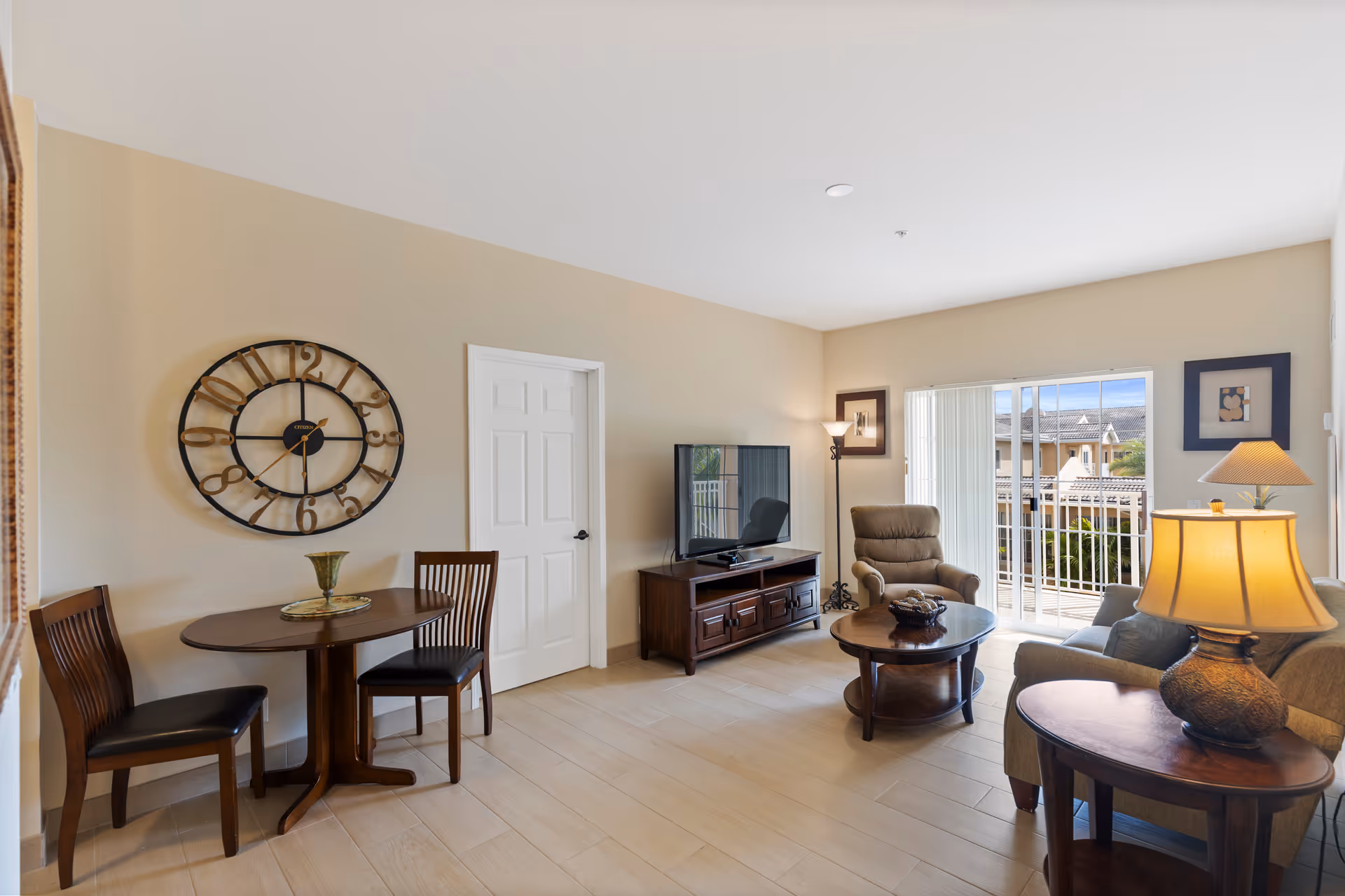 Sunlit living room with a sofa, armchair, coffee table, TV stand, small dining table, and sliding glass doors to a balcony.