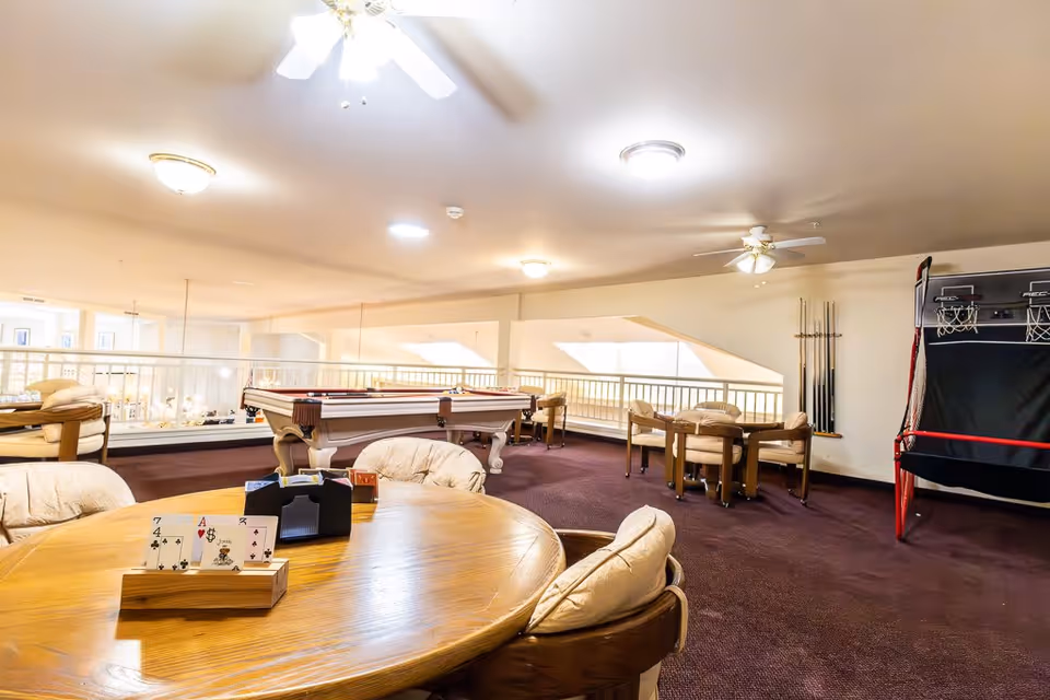 A spacious recreational room with a round wooden table and cushioned chairs in the foreground, a pool table in the middle, and a basketball arcade game on the right. The room has carpeted floors, ceiling fans, and bright overhead lighting, with a railing overlooking a lower level.