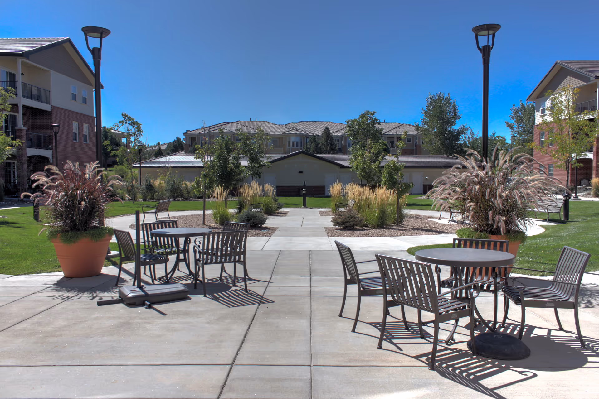 Outdoor patio area at Lincoln Meadows Senior Living with metal tables and chairs, large potted plants, lamp posts, and a walkway leading to residential buildings surrounded by grass and trees under a clear blue sky.