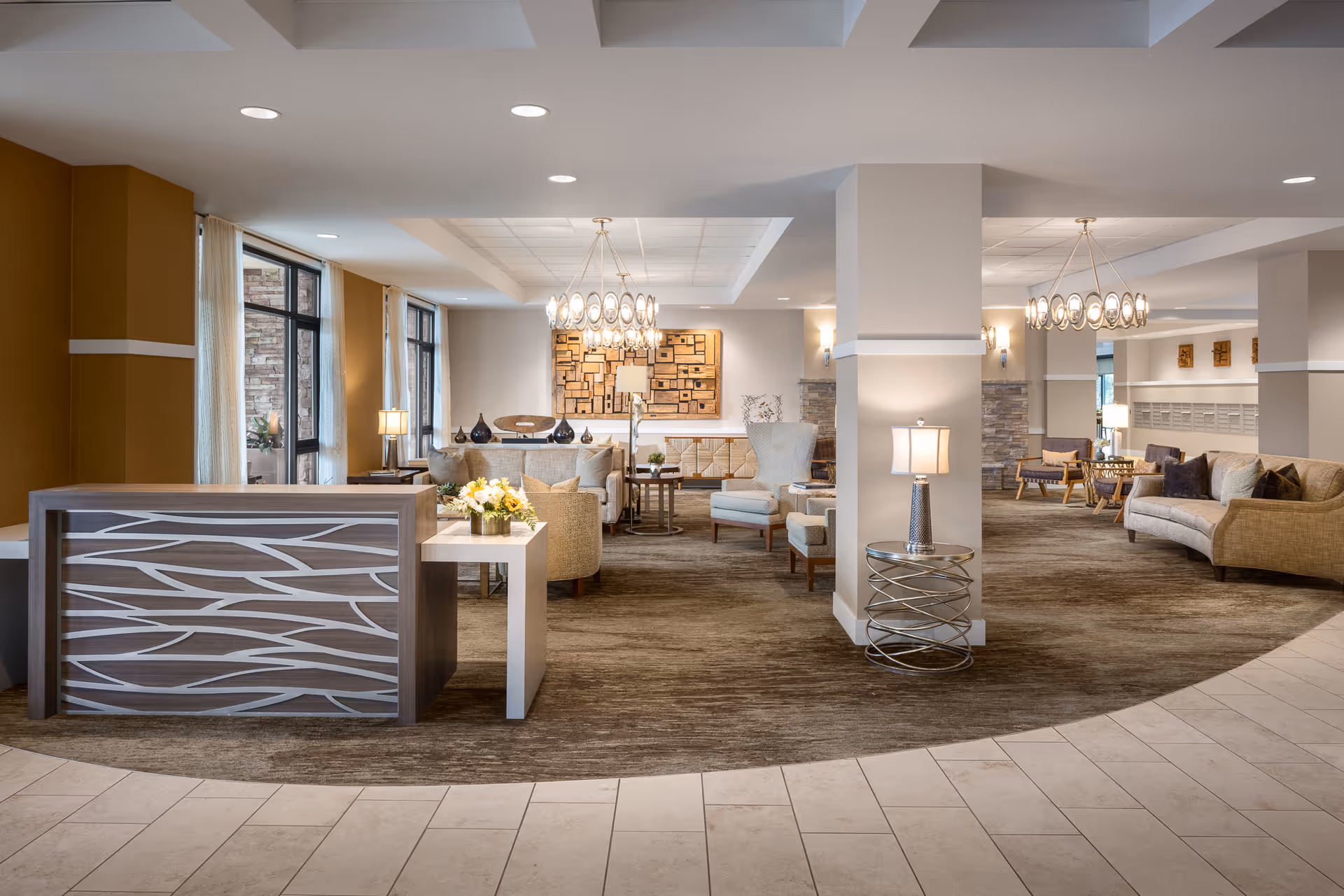 A spacious and well-lit senior living facility common area with modern decor, featuring a reception desk, comfortable seating arrangements including sofas and armchairs, decorative lamps, and large windows allowing natural light to enter.