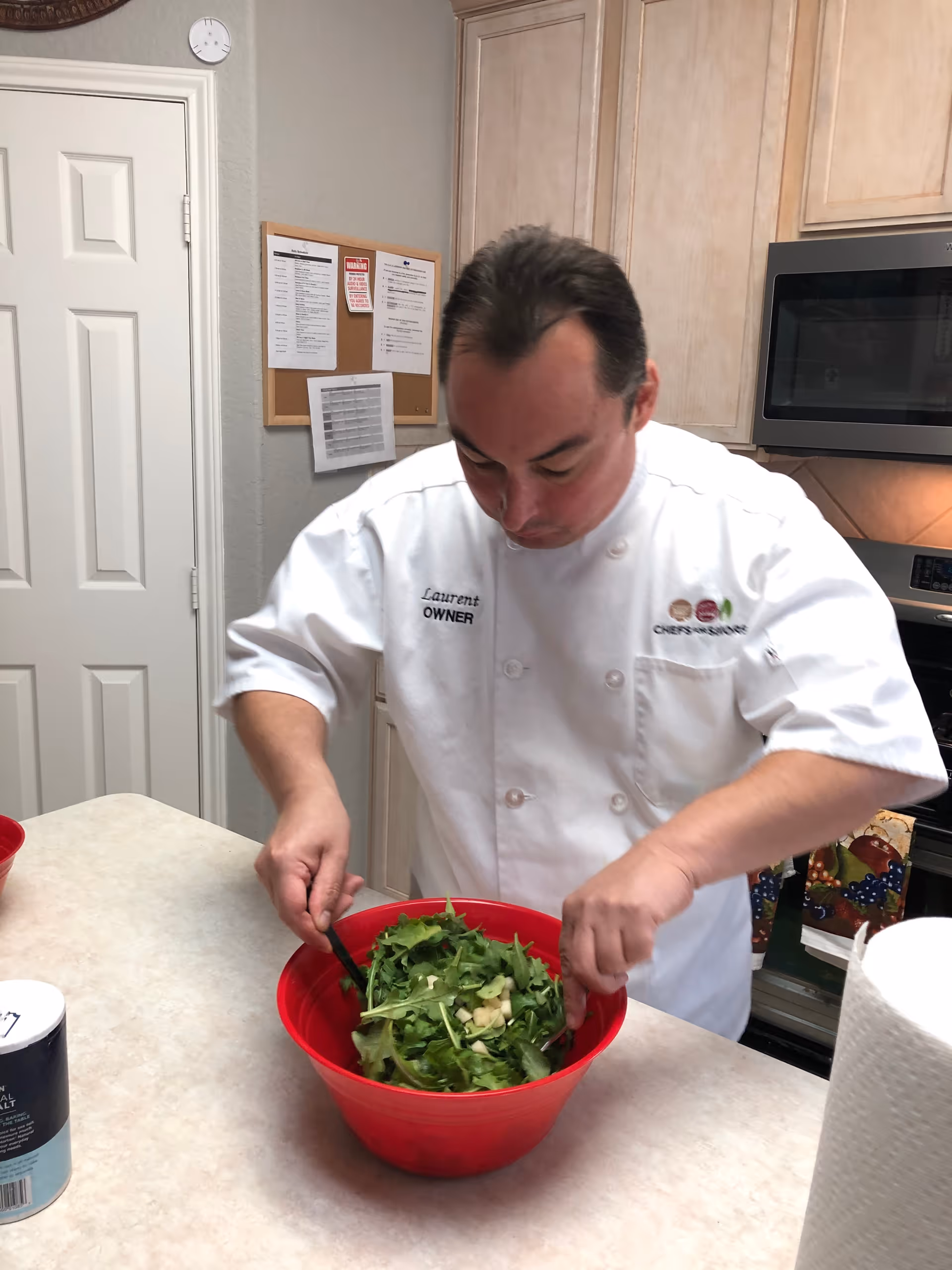 A man wearing a white chef's coat labeled 'Laurent OWNER' is preparing a salad in a red bowl on a kitchen counter. The kitchen has light-colored cabinets, a microwave, and a stove in the background. There is a paper towel roll and a container of salt on the counter.