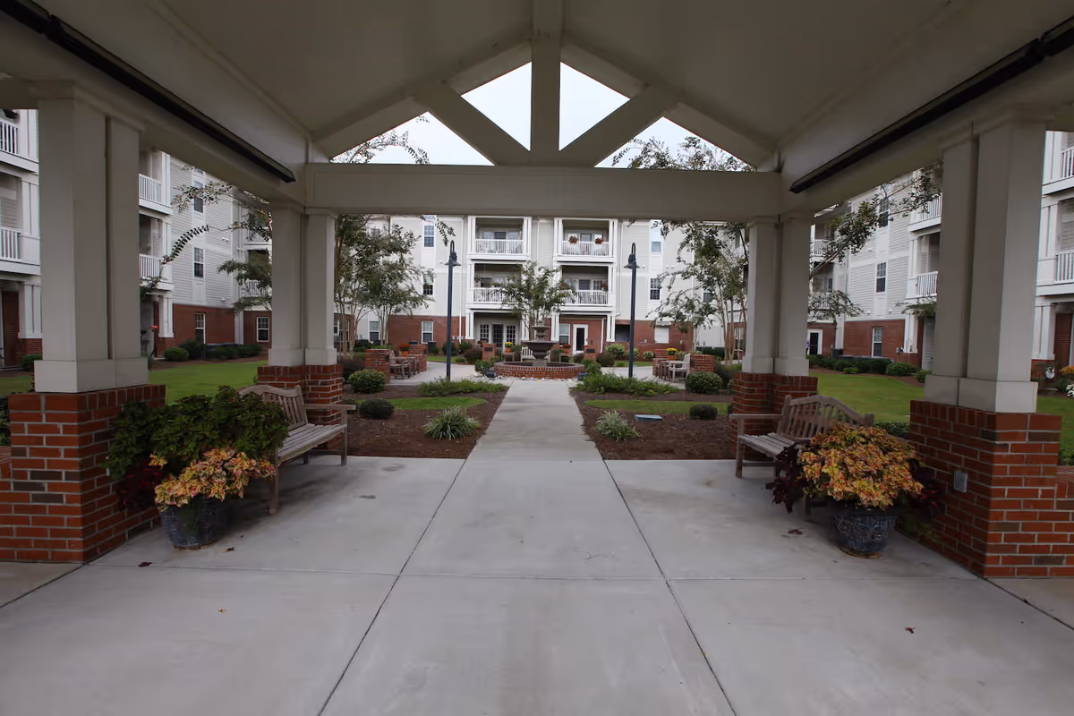 Covered walkway with benches and potted plants on either side, leading to a courtyard with trees, shrubs, and a multi-story residential building in the background.