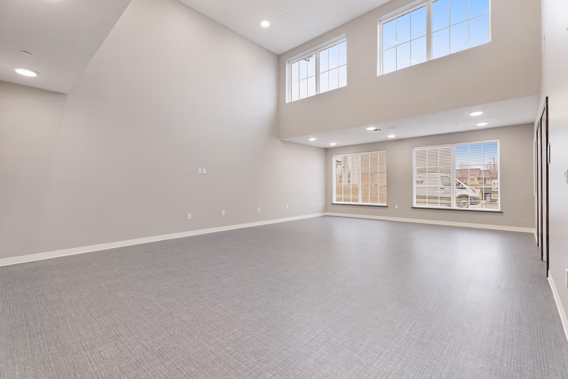 A spacious, empty room with high ceilings and large windows letting in natural light. The walls are painted light gray, and the floor has a textured gray finish. There are multiple recessed ceiling lights and electrical outlets along the walls.