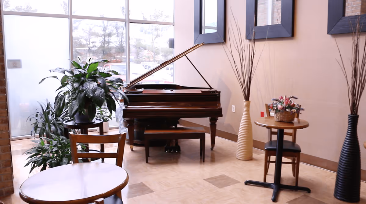 Interior view of a common area with a grand piano, several potted plants, small round tables with chairs, and tall decorative vases with branches. Large windows allow natural light to fill the space.