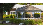 A white wooden gazebo with a shingled roof situated in a garden area with green bushes, flowers, and trees. A building with white-framed windows is visible in the background.