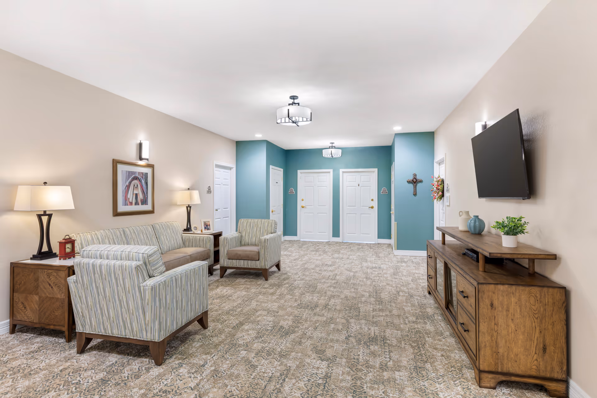 A spacious, well-lit common area with beige walls and a patterned carpet. The room features a striped sofa and two matching armchairs arranged around a wooden side table with lamps. On the right wall, there is a mounted flat-screen TV above a wooden cabinet decorated with small plants and vases. The far wall is painted teal and has four white doors with gold handles. The ceiling has two modern light fixtures, and the room has a calm, welcoming atmosphere.