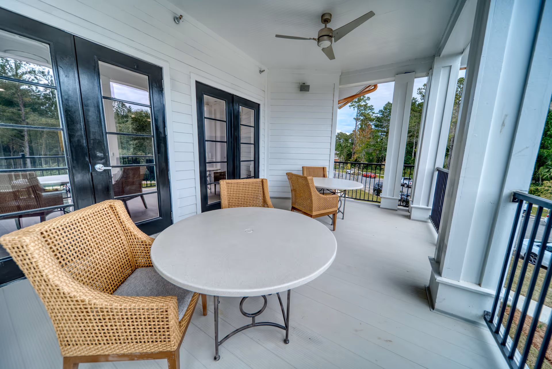 Covered outdoor balcony area with two round tables and four wicker chairs. The balcony has white walls, black-framed glass doors, a ceiling fan, and black railings. Trees and parked cars are visible in the background.