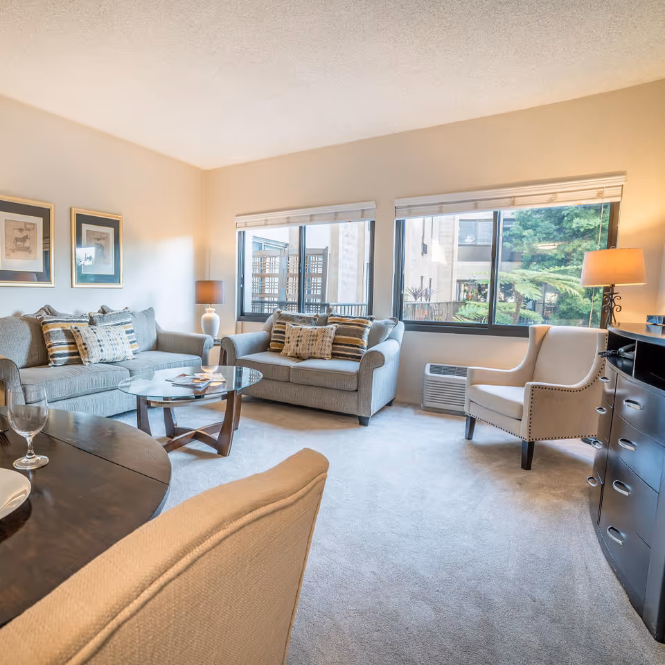 Sunlit living room with two gray sofas, an armchair, glass coffee table, and large windows overlooking greenery.