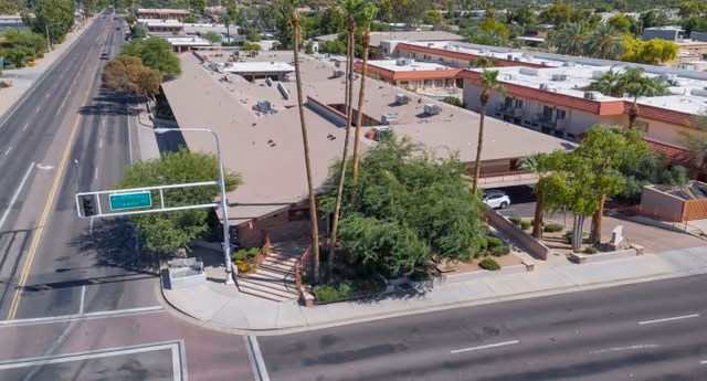 Aerial view of a single-story building complex with a beige roof, surrounded by trees and landscaping, located at a street intersection with traffic lights and road signs. The area appears to be in a suburban neighborhood with other buildings and greenery nearby.