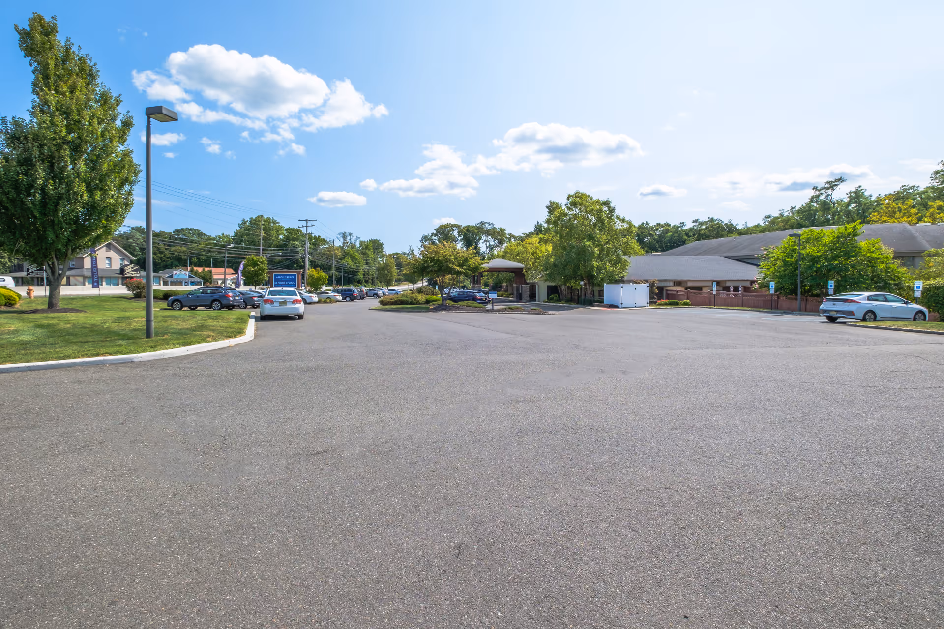 Wide view of a parking lot outside Arbor Terrace Middletown senior living facility on a sunny day with a few cars parked, trees, and a clear blue sky with some clouds.