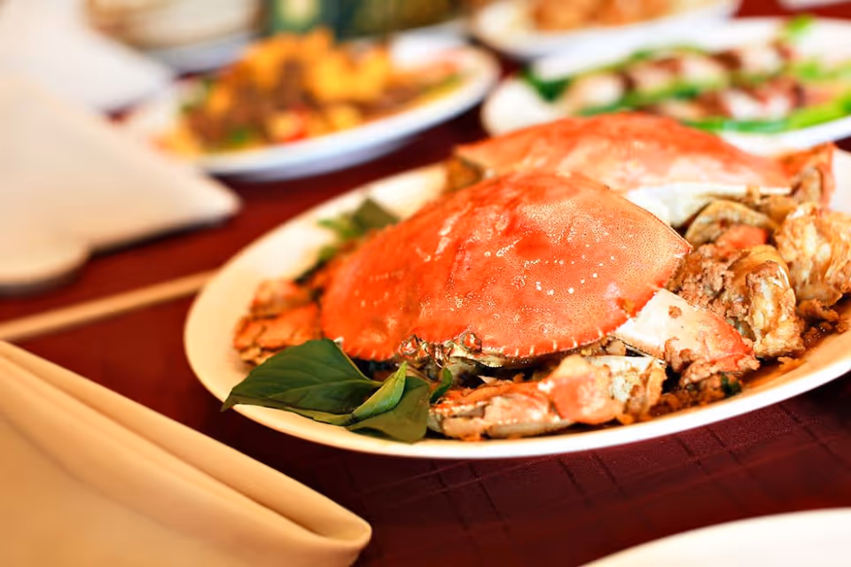 Close-up of a plate with cooked whole crabs garnished with green leaves on a table with a maroon tablecloth, with other plates of food blurred in the background.
