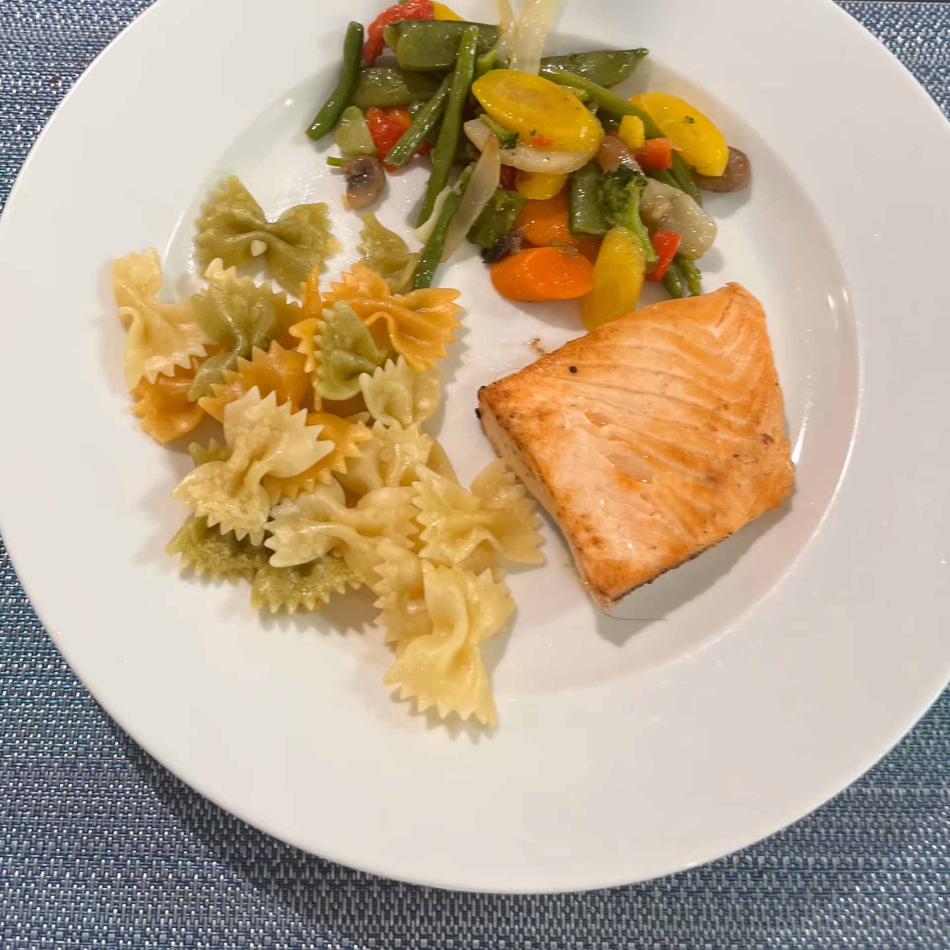 A white plate with a serving of cooked bow-tie pasta in three colors, a piece of grilled salmon, and a portion of mixed sautéed vegetables including green beans, yellow squash, carrots, mushrooms, and red bell peppers on a blue woven placemat.