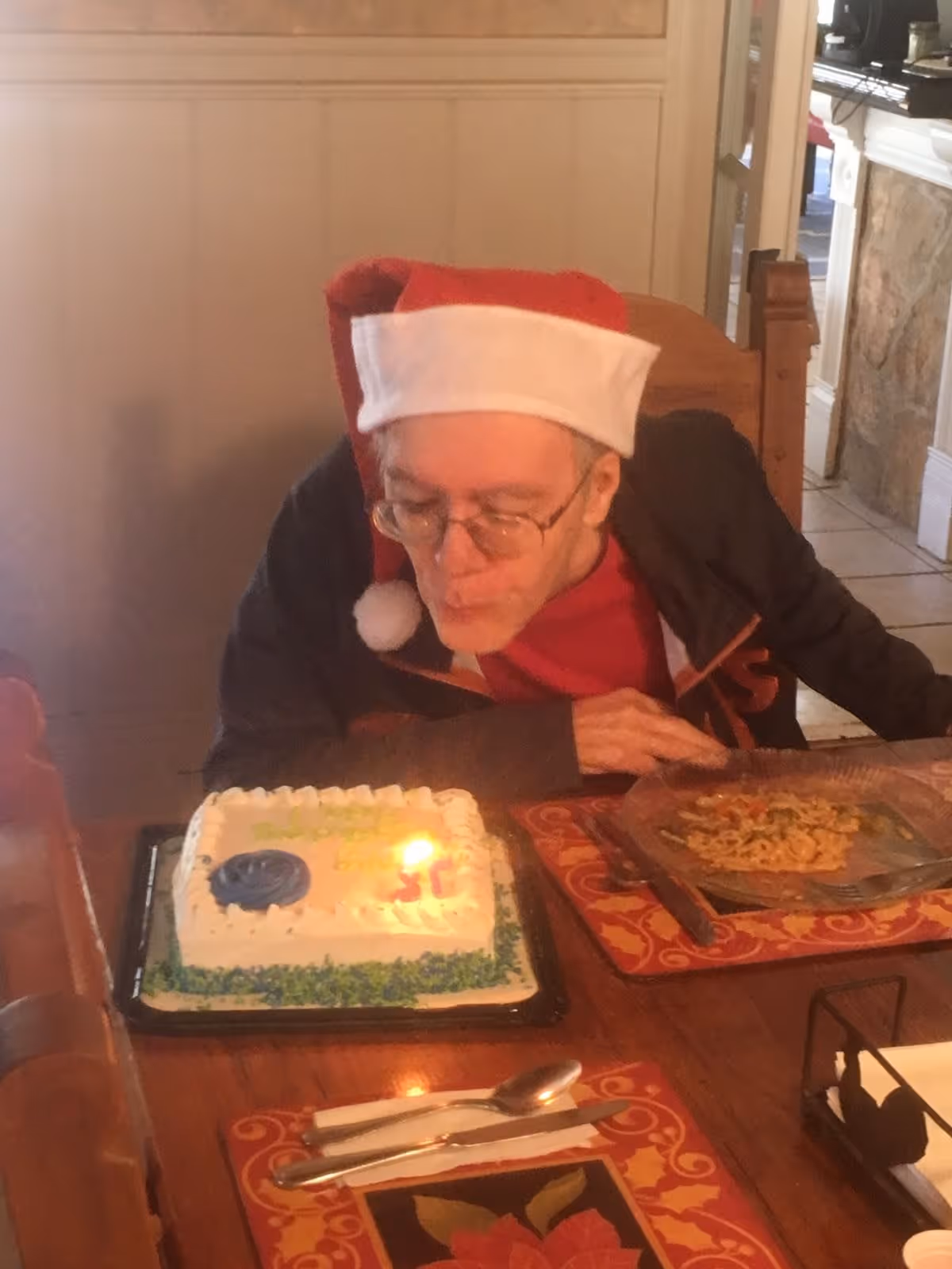 A person wearing a Santa hat leans over a dining table to blow out a candle on a rectangular birthday cake, with a plate of food and place settings nearby.
