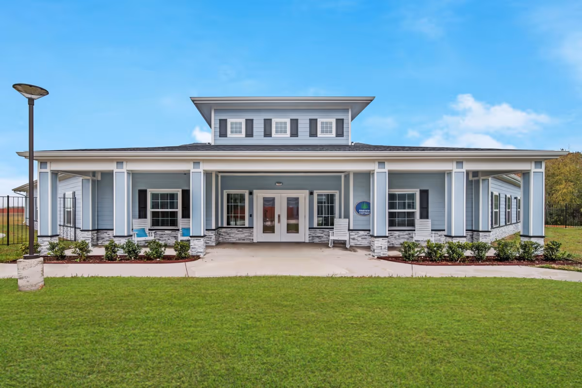Front exterior view of a single-story building with light blue siding, white trim, and a covered porch with white rocking chairs. The building is surrounded by a well-maintained lawn and small shrubs, under a clear blue sky.