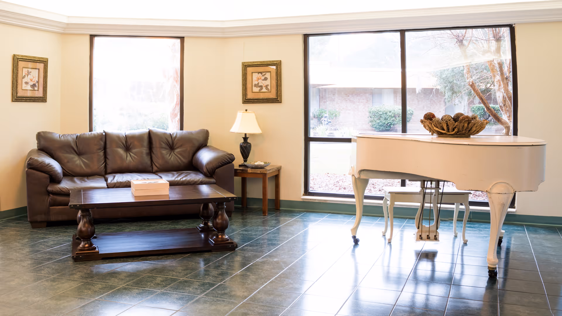 A bright living room with a dark brown leather sofa, a wooden coffee table with a box on it, a side table with a lamp, two framed floral pictures on the wall, and a white grand piano near large windows showing an outdoor garden.