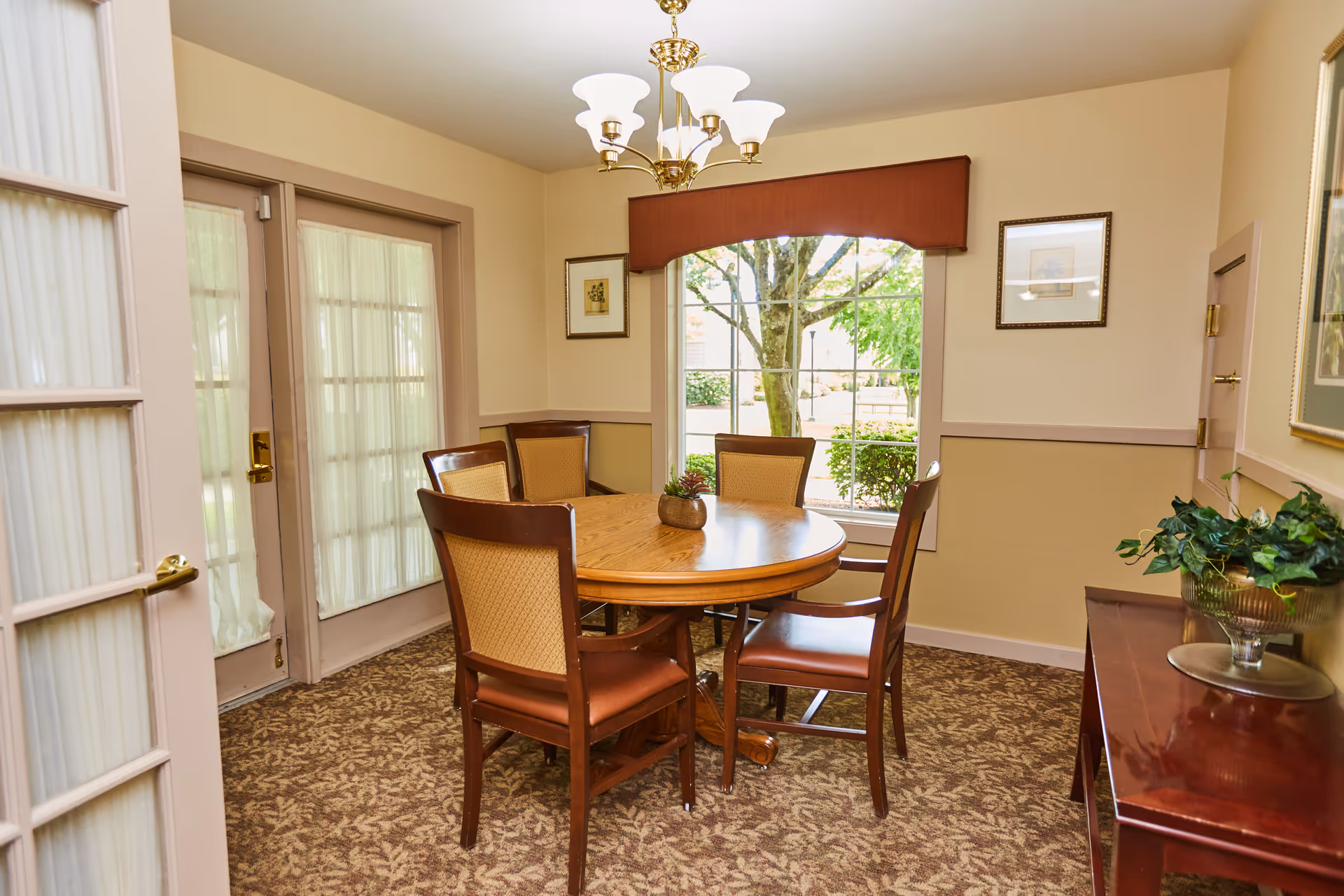 A cozy dining area with a round wooden table surrounded by five wooden chairs with cushioned seats and backs. The room has beige walls and carpet with a leafy pattern. A large window with a red valance shows a view of greenery outside. French doors with sheer curtains are on the left side. A wooden side table with a decorative plant is on the right, and a chandelier with five lights hangs from the ceiling.