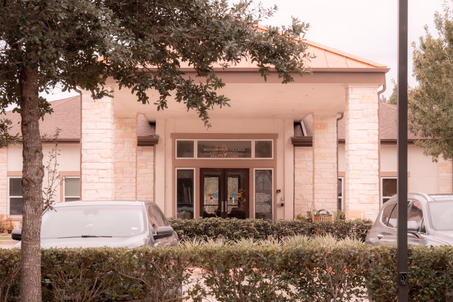 Front entrance of Woodhaven Village assisted living building with stone columns, double glass doors, parked cars, and hedges.