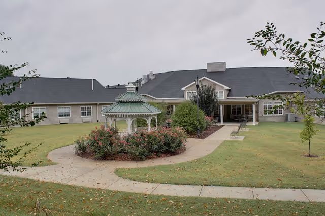 Exterior view of a single-story building with a gray roof and beige siding, featuring a central entrance with a covered porch. In front of the building, there is a circular garden area with a white gazebo surrounded by bushes and flowers. The lawn is green with a few small trees and a concrete pathway leading to the entrance and around the garden.