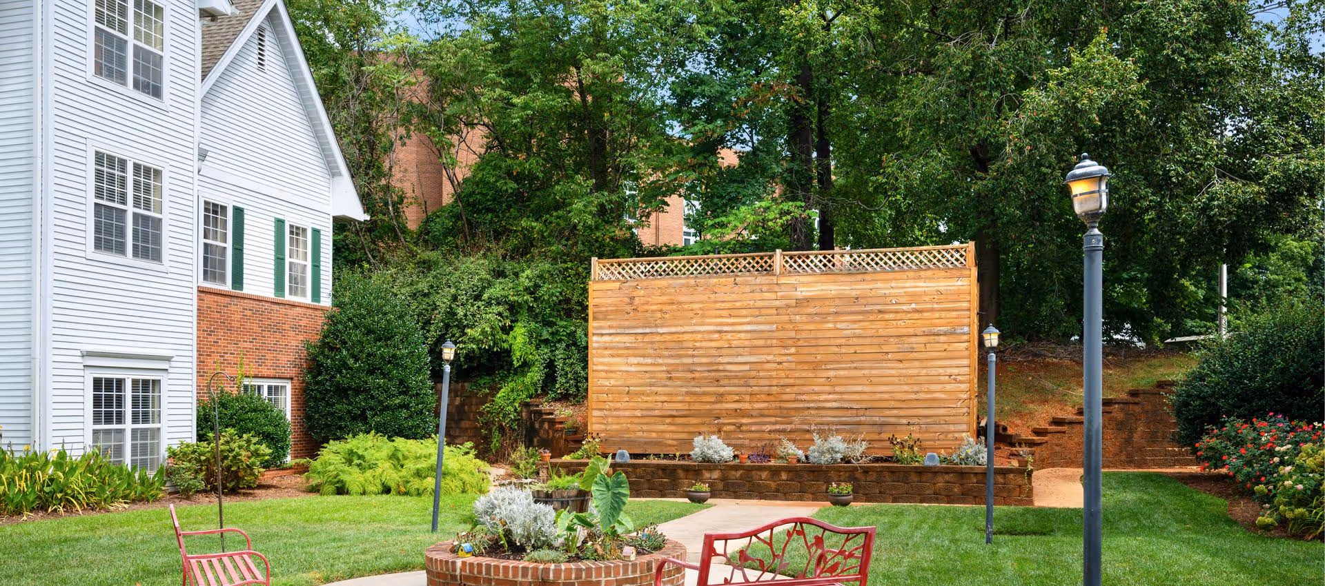 Outdoor garden area with green grass, a circular brick planter with plants, red metal benches, lamp posts, and a wooden privacy fence. A white and brick building is visible on the left side, with trees and shrubs surrounding the area.