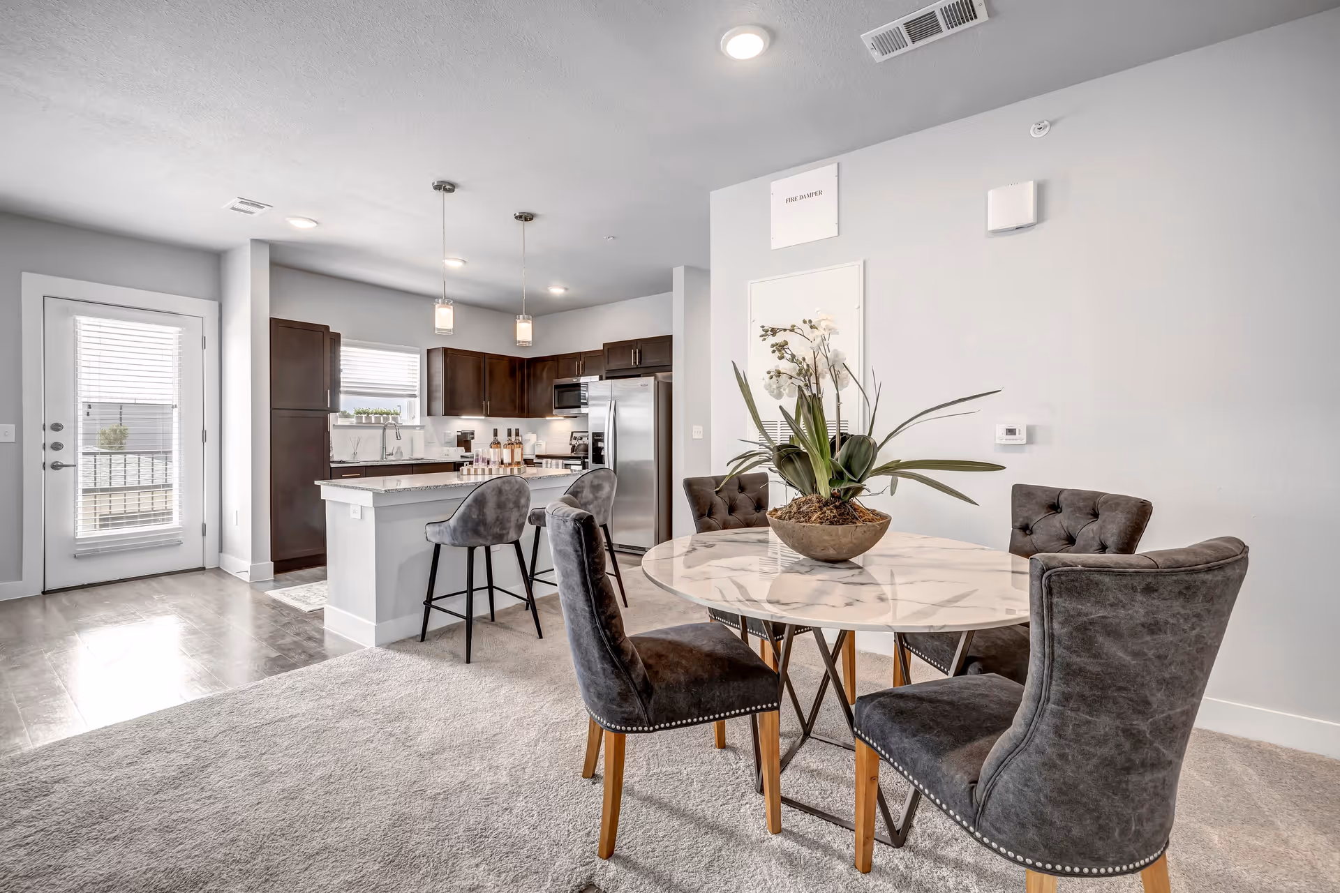 A modern open-concept kitchen and dining area featuring a round marble dining table with four dark upholstered chairs. The kitchen has dark wood cabinets, a stainless steel refrigerator, a kitchen island with two gray bar stools, and pendant lights hanging above the island. There is a door with blinds leading outside and a window above the kitchen sink.