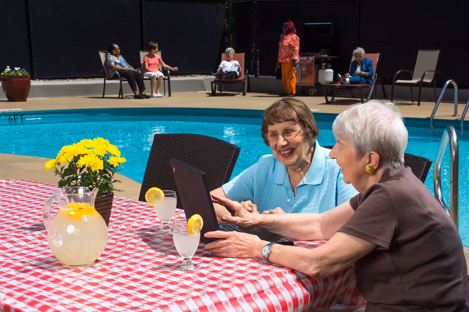 Two elderly women sitting at a table with a red and white checkered tablecloth near a swimming pool, looking at a laptop and smiling. The table has a pitcher of lemonade, two glasses with lemon slices, and a pot of yellow flowers. In the background, other elderly people are sitting on chairs and lounge chairs by the pool, with one person standing near a grill.