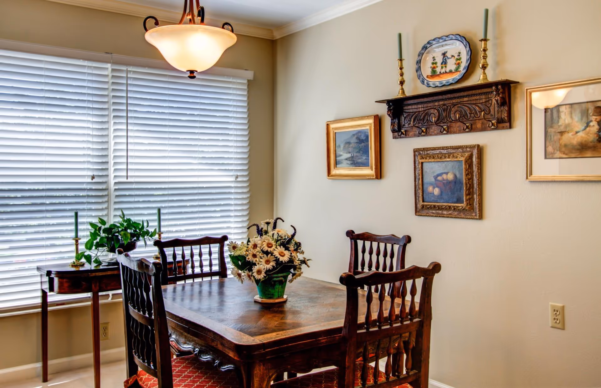 A cozy dining room with a wooden table and four matching chairs. A floral arrangement in a green vase is centered on the table. The room has large windows with white blinds, a small side table with a plant and two candlesticks, and three framed paintings on the beige walls. A decorative wooden shelf with two candlesticks and a plate is mounted above the paintings.