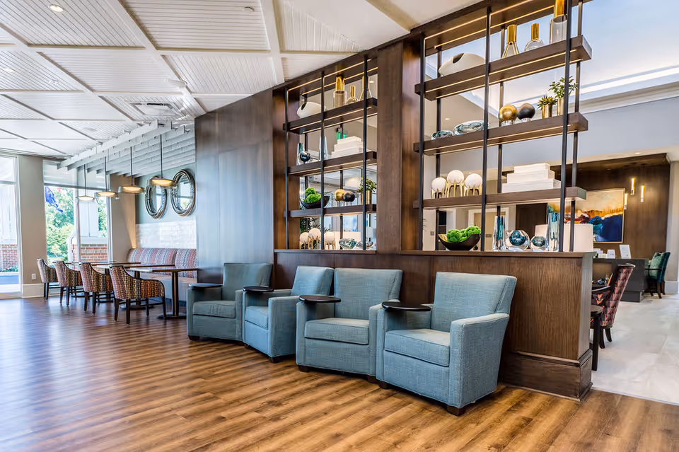 A bright senior living common area with blue armchairs in front of decorative shelving and a dining area with tables and chairs in the background.