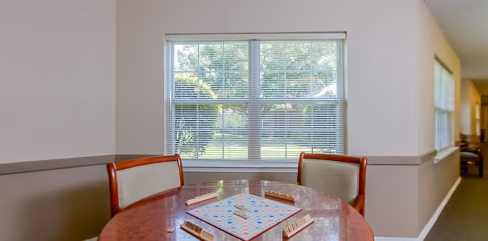 Round wooden game table with a Scrabble board and two chairs set in front of a large window overlooking a lawn.