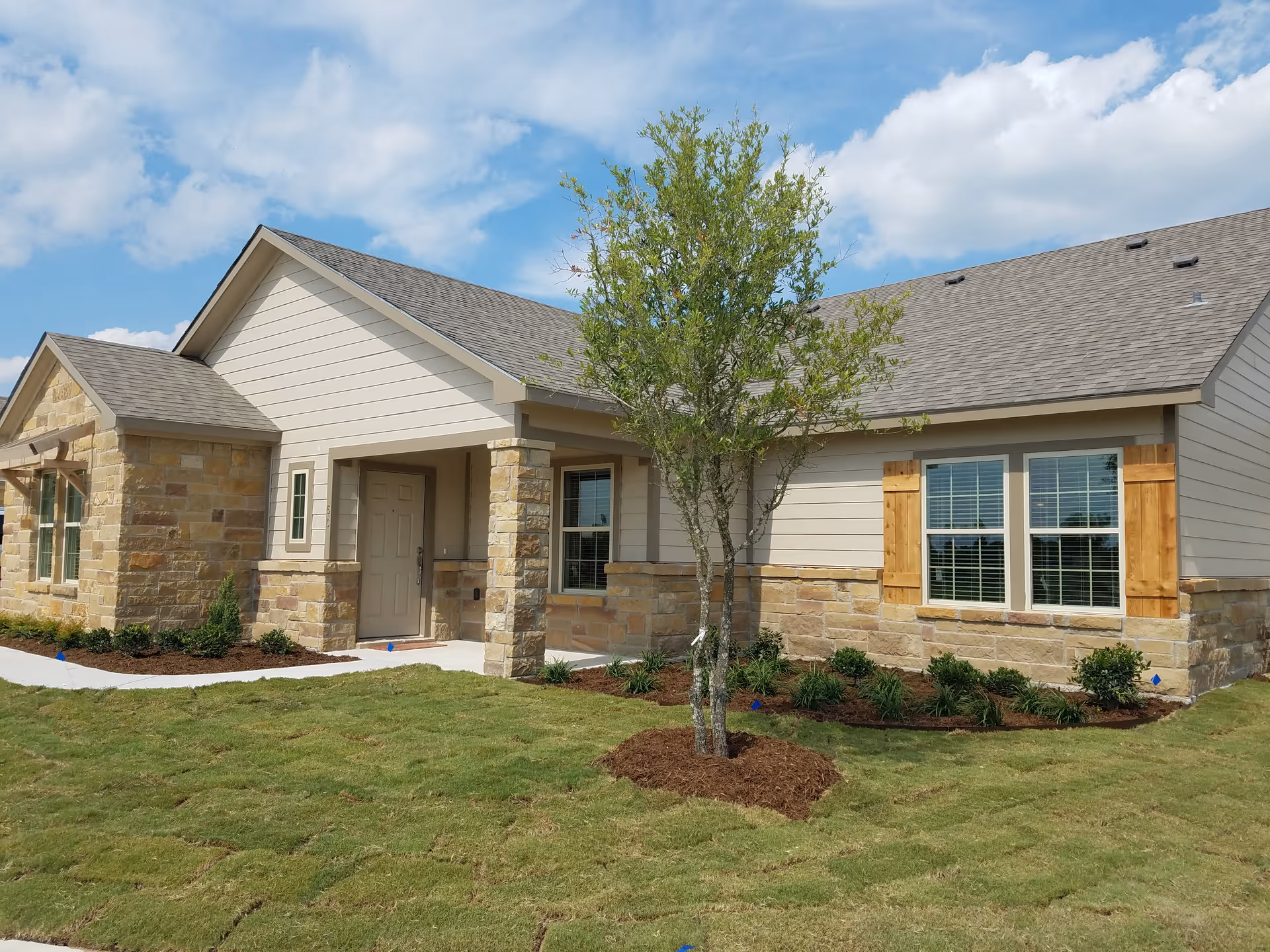 Single-story cottage-style building with stone and siding facade, front porch, windows with wooden shutters, a small tree, and a freshly sodded lawn.