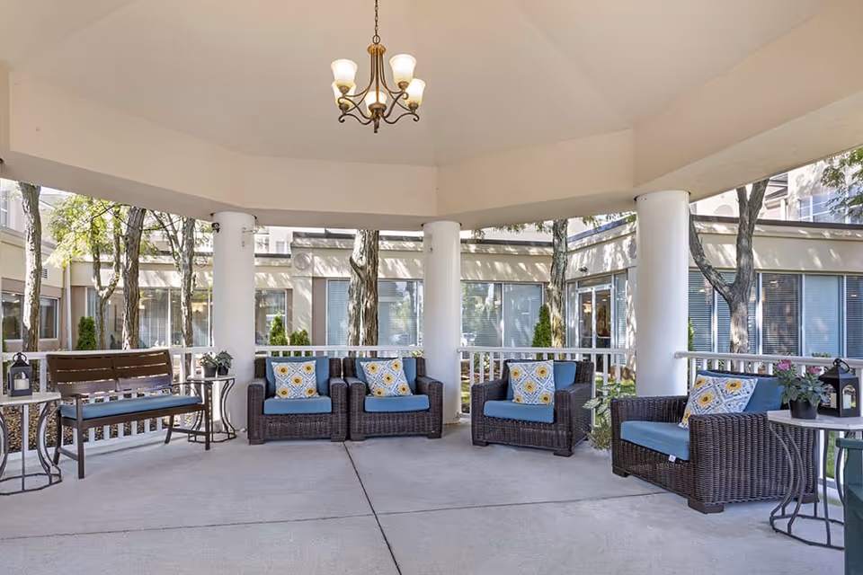 Covered outdoor seating area with wicker chairs and blue cushions, decorative pillows, a wooden bench, small side tables with plants, white columns, and a hanging chandelier light fixture. The area is surrounded by windows and trees.