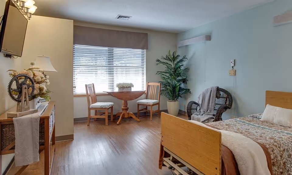 Furnished senior bedroom with a hospital-style bed, wicker chair, small round table and two chairs by a large window, TV and potted plant.