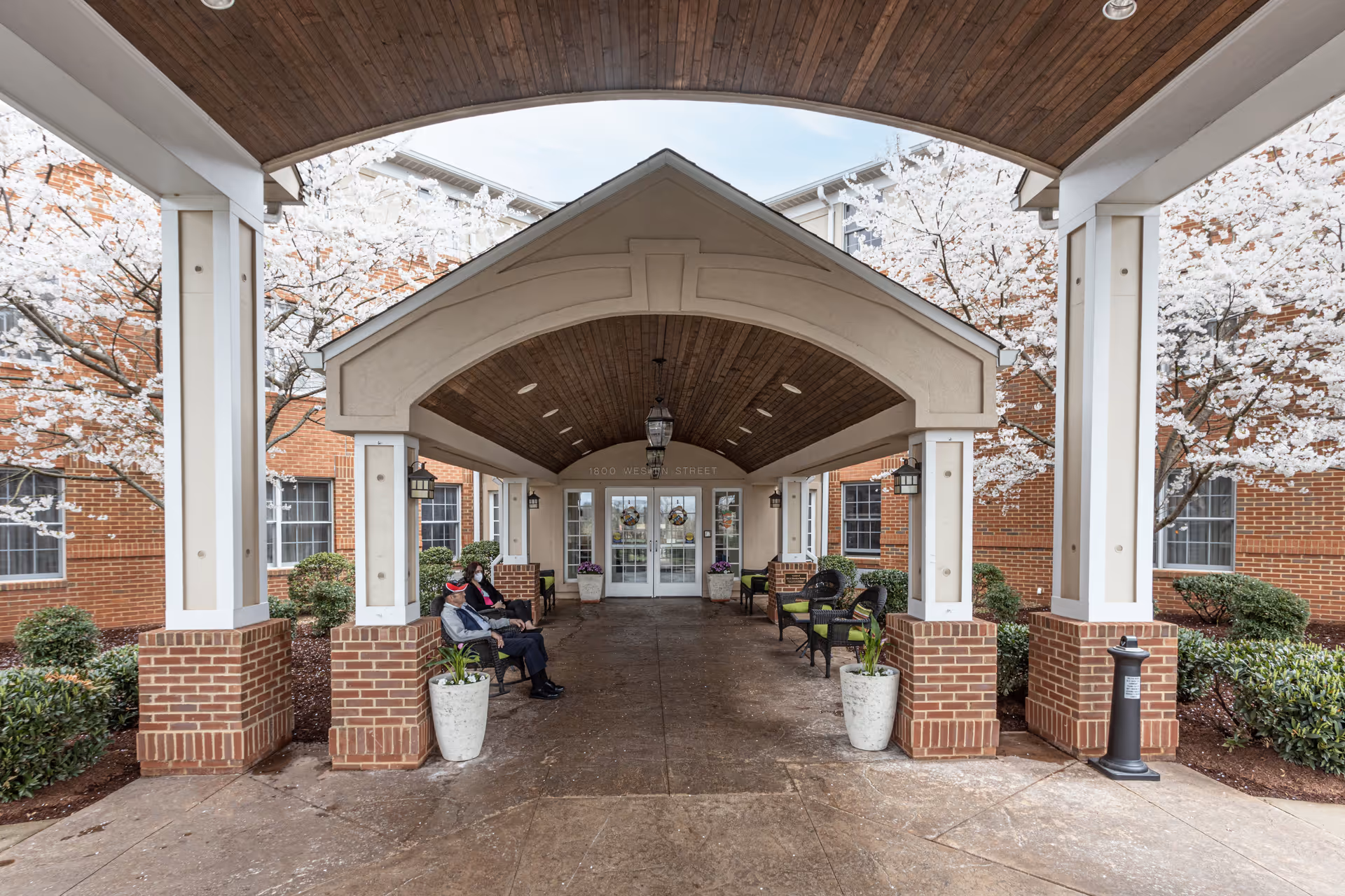 Covered entrance portico of a brick senior living building with benches, planters, blooming trees, and two people seated.