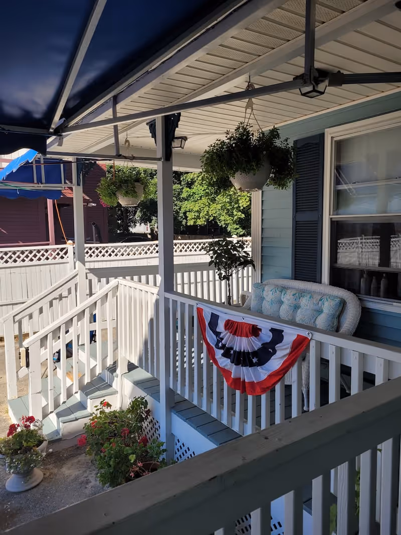 A covered porch with white railings and steps leading down to a garden area with potted plants. The porch has hanging plants and a cushioned wicker bench with a patriotic red, white, and blue bunting decoration on the railing. The house exterior is light blue with dark blue shutters and a window.