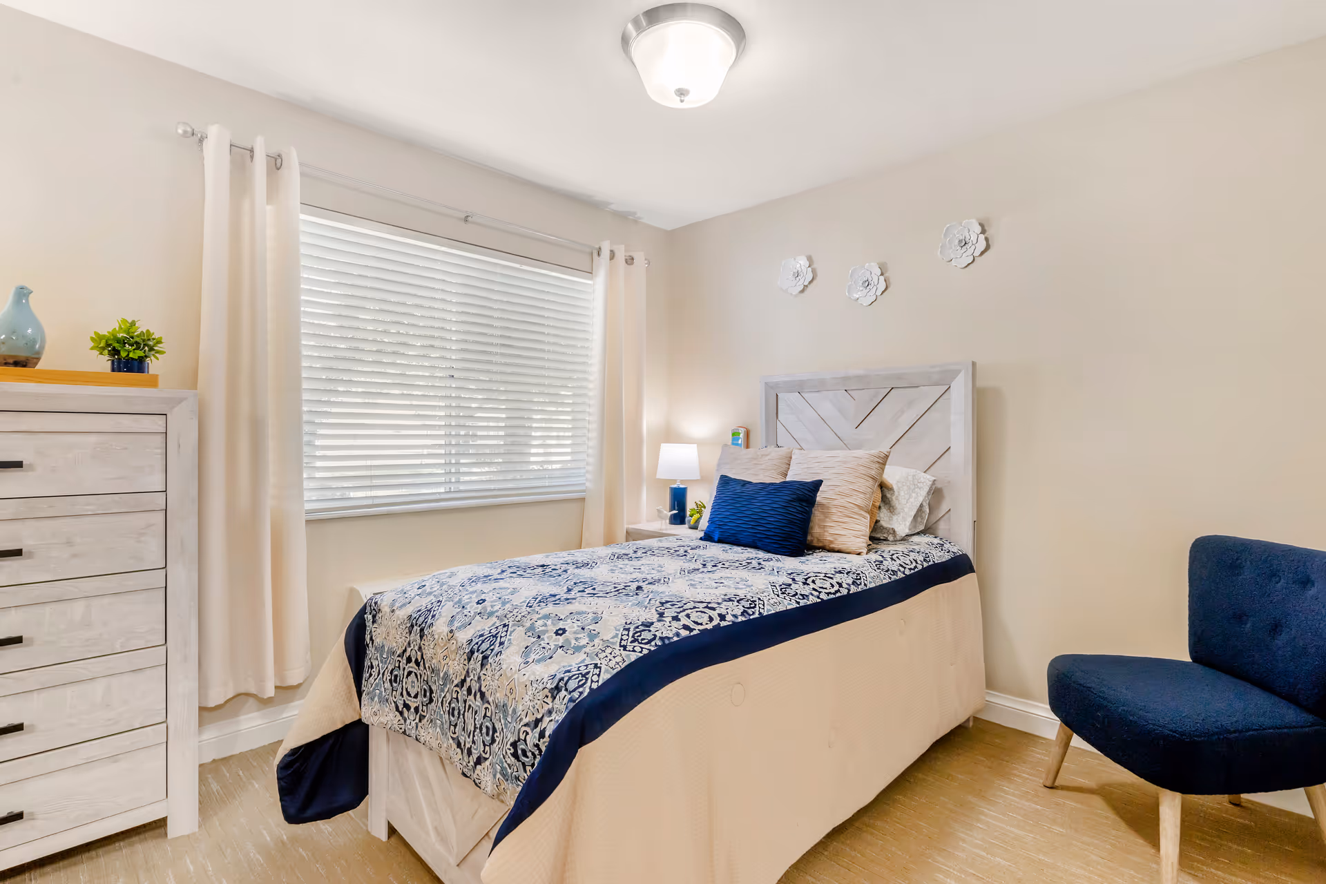 Bright bedroom with a single bed featuring blue patterned bedding, a dresser, a window with blinds and curtains, a bedside lamp, and a blue chair.