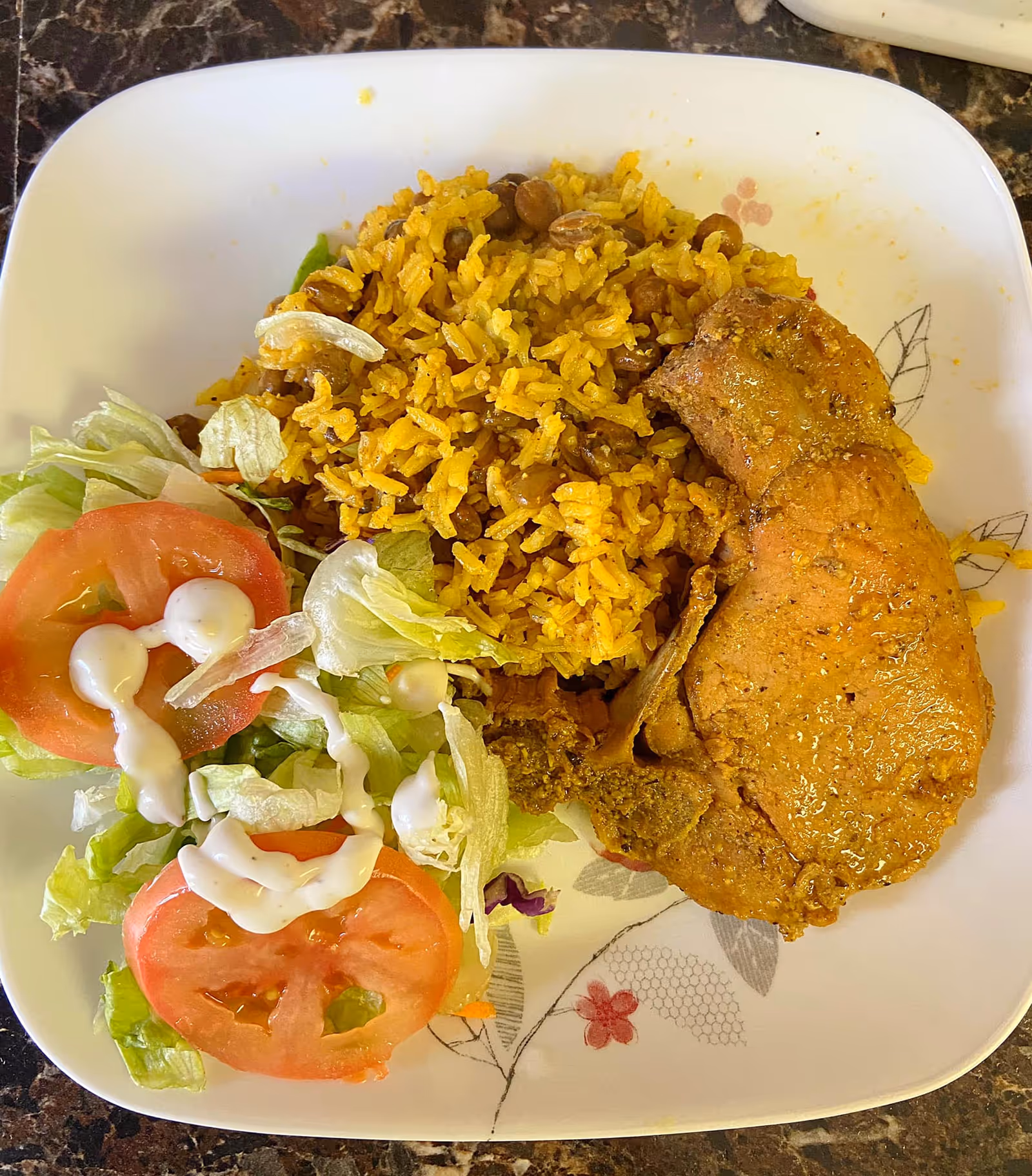 Plate with seasoned chicken, yellow rice with beans, and a side salad of lettuce and tomato topped with dressing.