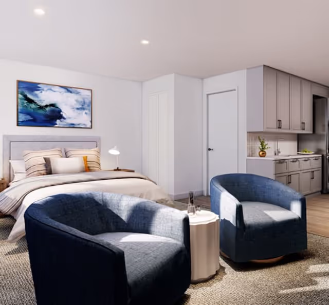 Studio-style interior showing a bed, two blue armchairs around a small side table, and a kitchenette along one wall.