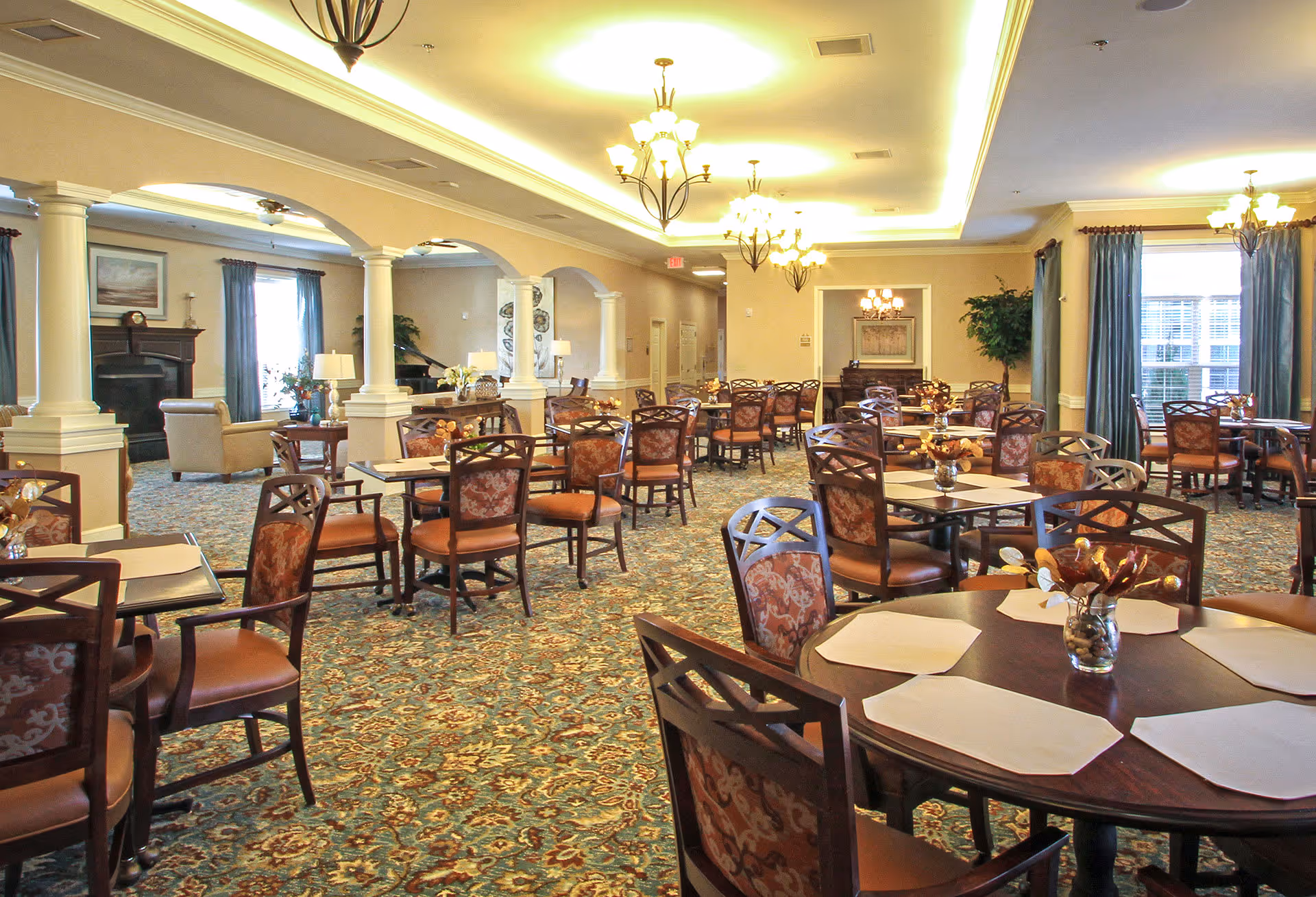 A spacious dining room with multiple round and square tables set with placemats and small floral centerpieces. The room features patterned carpet, beige walls, large windows with blue curtains, and elegant chandeliers hanging from the ceiling. There is a seating area with armchairs and a fireplace visible in the background.
