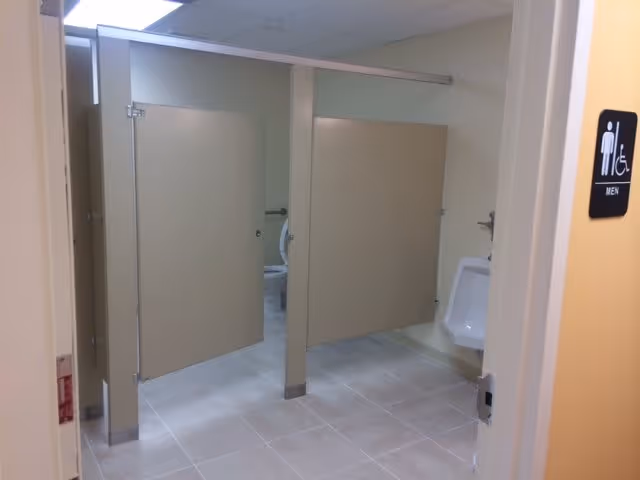 Public men's restroom with beige stall partitions, a urinal, tiled floor, and an accessible men's sign on the doorframe.