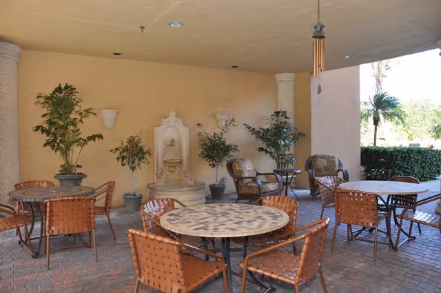 Covered outdoor seating area with round mosaic tables, woven chairs, potted plants, a wall fountain and a wind chime.