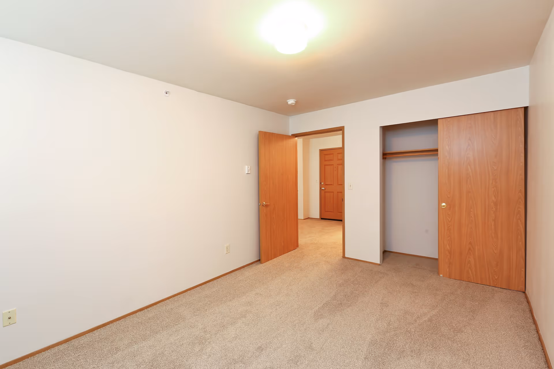 Empty bedroom with beige carpet, white walls, a wooden door open to a hallway, and a closet with sliding wooden doors.