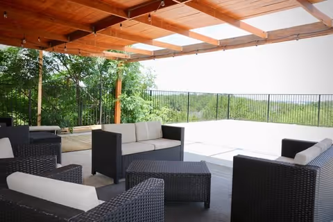 Outdoor covered patio area with black wicker furniture including sofas and chairs with white cushions, overlooking a fenced area with greenery and trees in the background.