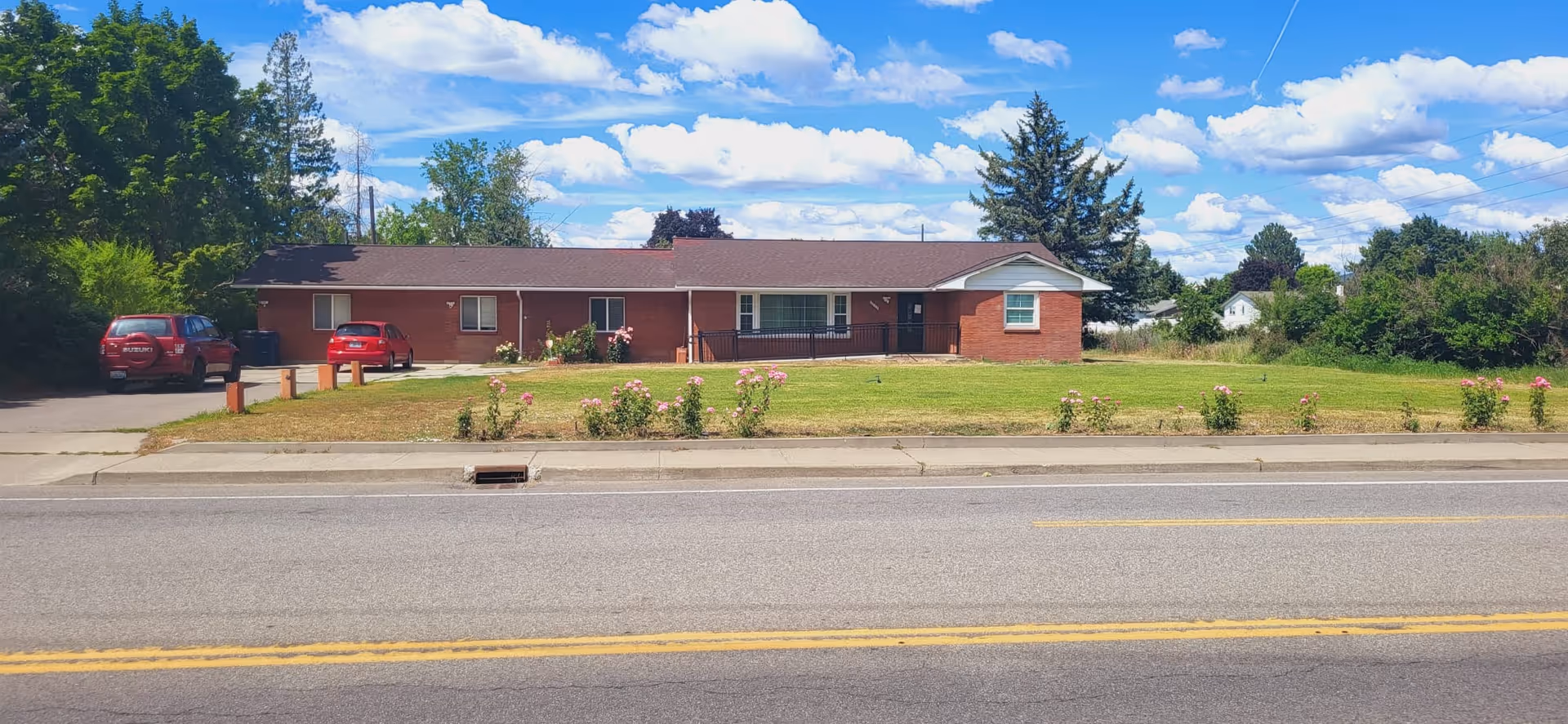 Single-story brick ranch house with a large front lawn, driveway and two parked cars under a blue sky.