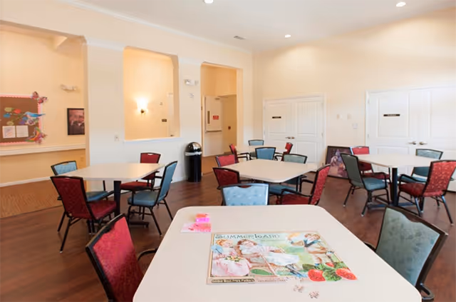 Communal dining/activity room with several square tables and red and blue chairs and a puzzle on the nearest table.