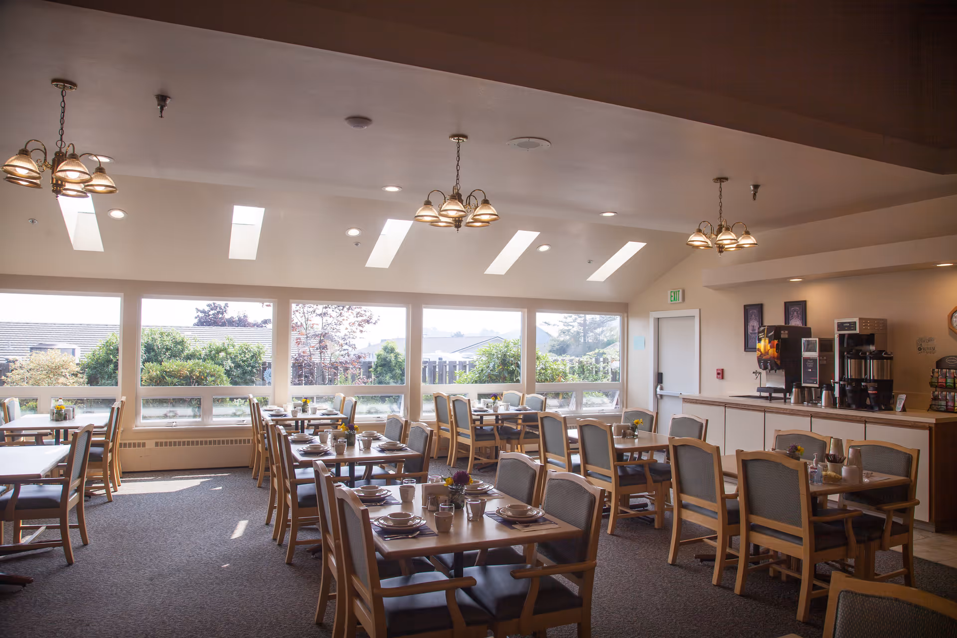 A bright dining room with multiple wooden tables and chairs set with plates, cups, and utensils. Large windows along one wall let in natural light and offer views of greenery outside. Ceiling lights and skylights illuminate the room. A beverage station with coffee and juice dispensers is visible in the background.