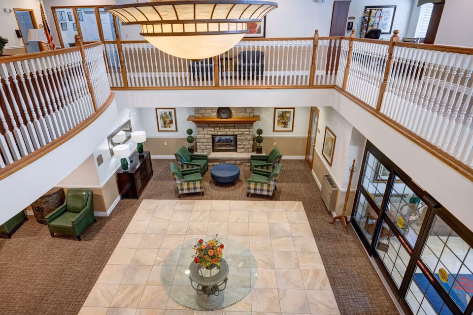 View from above of a spacious senior living facility lobby with a tiled floor area featuring a round glass table with a floral arrangement. Surrounding the tiled area is carpeted flooring with green armchairs and a stone fireplace with framed artwork on the walls. The upper level has white railings with wooden handrails, and there is a large ceiling light fixture hanging above.