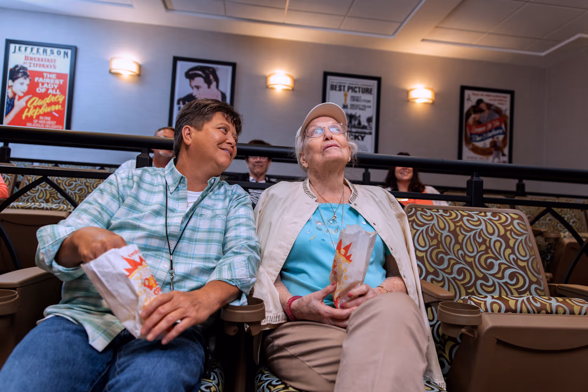 Two elderly women sitting in a theater-style room with patterned seats, holding popcorn bags. One woman is wearing a light green plaid shirt and smiling at the other woman, who is wearing a light blue shirt, beige jacket, and a white cap. Movie posters are visible on the wall behind them.
