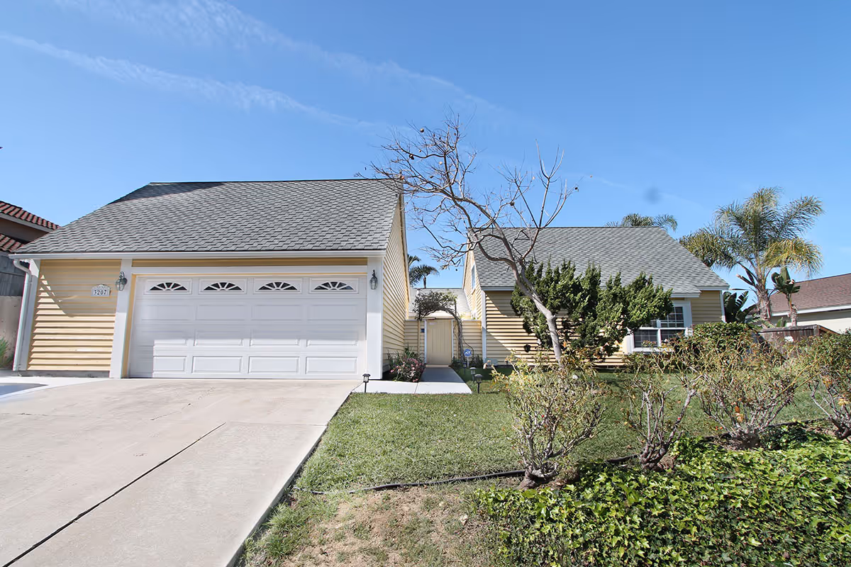 Front exterior view of a single-story yellow house with a gray shingled roof, a two-car garage, a driveway, a small lawn, and some bushes and trees in the yard under a clear blue sky.
