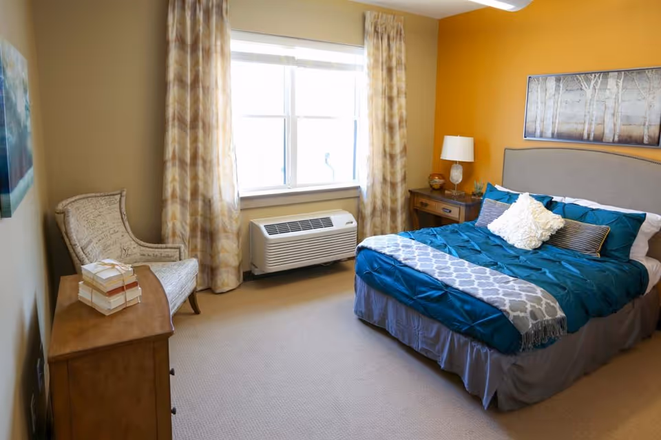 A cozy bedroom with a bed dressed in teal and gray bedding, a white decorative pillow, a wooden nightstand with a lamp and decorative items, a cushioned armchair, a wooden dresser with stacked books, beige carpet, and a window with patterned curtains letting in natural light.
