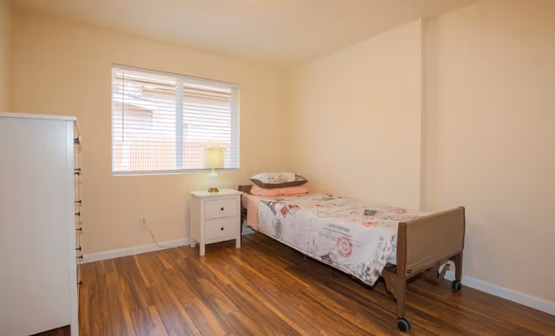 A simple bedroom with a single bed on wheels covered with a patterned bedspread, a white nightstand with a lamp, a window with blinds, and a white dresser on the left side. The room has wooden flooring and cream-colored walls.