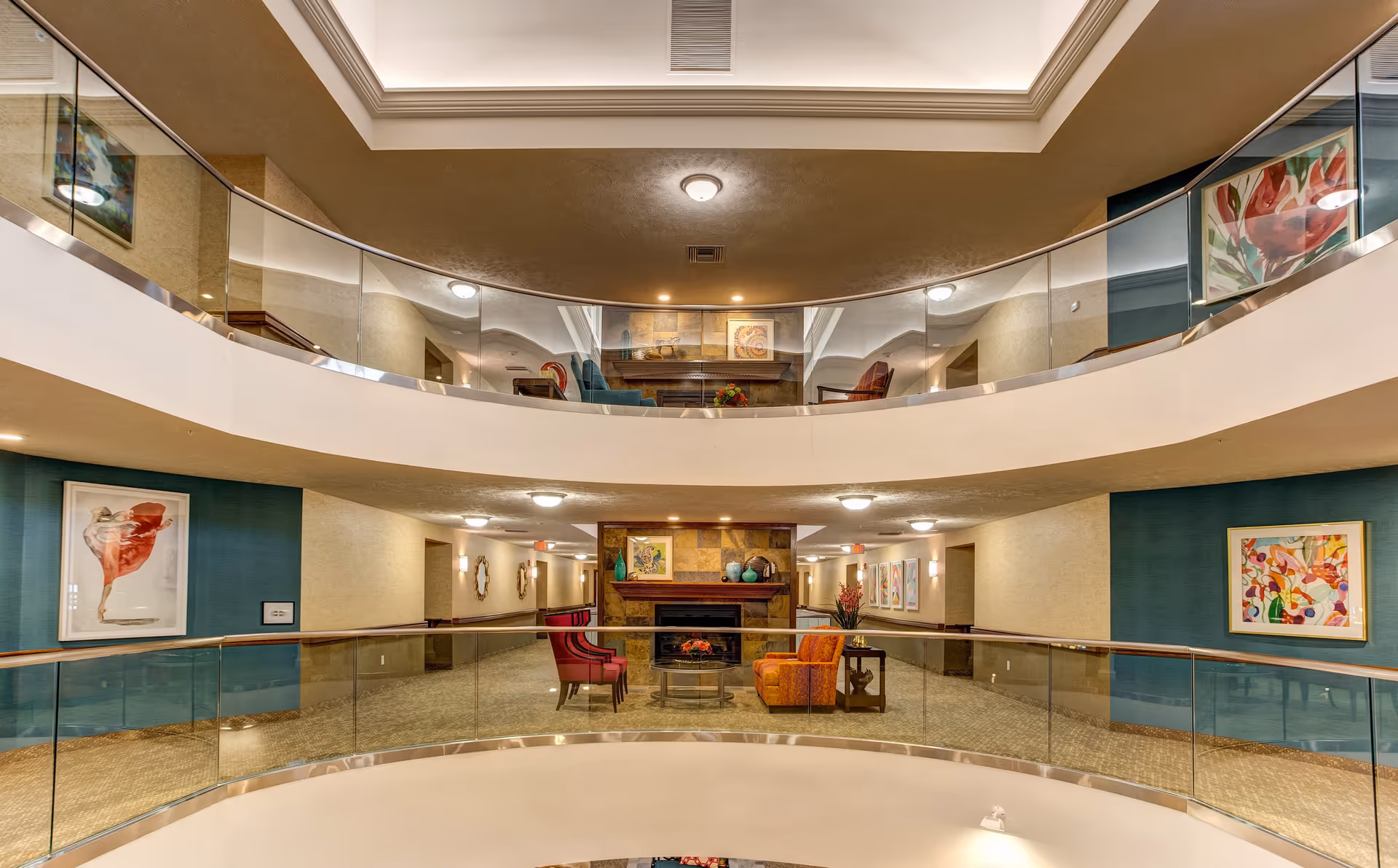 Interior view of a senior living facility with two levels visible. The lower level features a seating area with colorful chairs and a fireplace, surrounded by glass railings. The upper level has additional seating and artwork on the walls. The space is well-lit with ceiling lights and decorated with framed paintings.